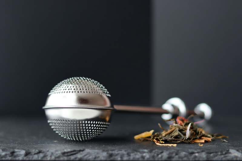 Close-up of hands using a stainless steel tea infuser over a glass teapot filled with herbal tea